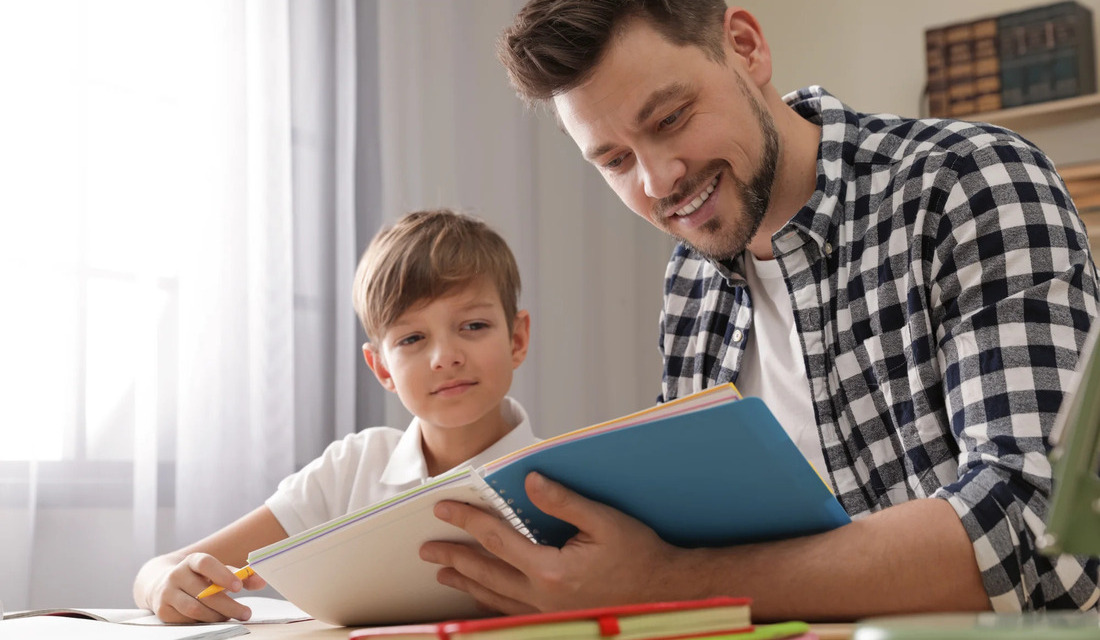 A man and a boy are at a table, both looking at a book, highlighting a bonding moment through reading.