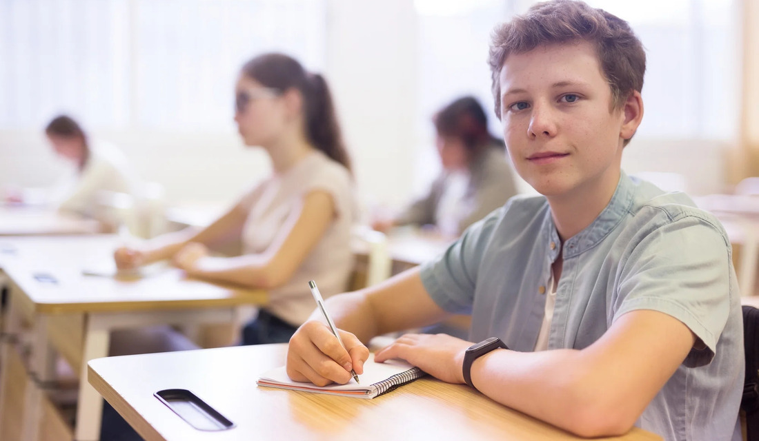 A young man seated at a classroom desk, focused on his work with books in front of him.