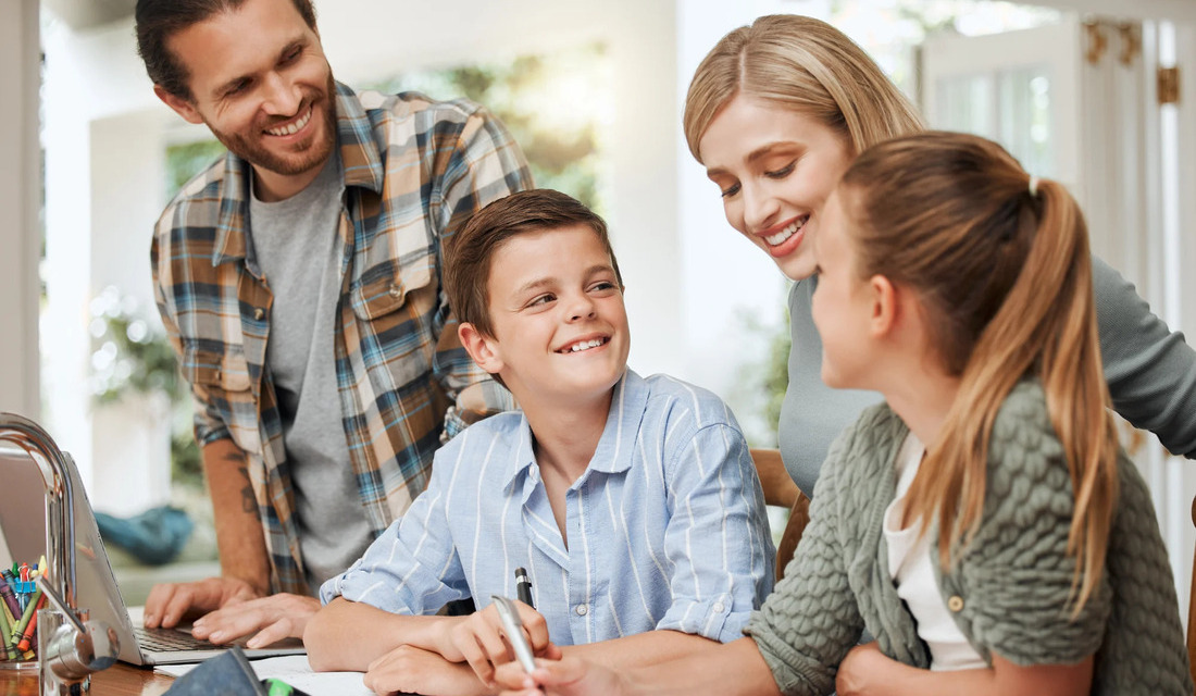 A family of four sits around a table, engaging with a laptop, sharing smiles and conversation.