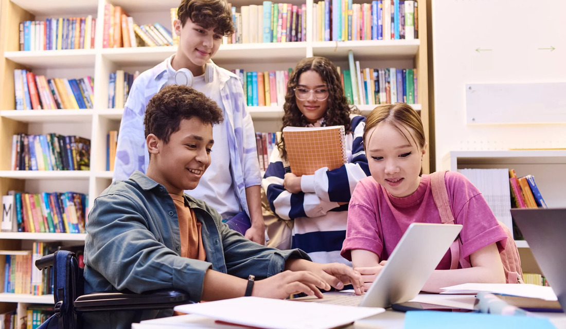 A group of students seated at tables in a library, focused on their laptops while studying and collaborating.