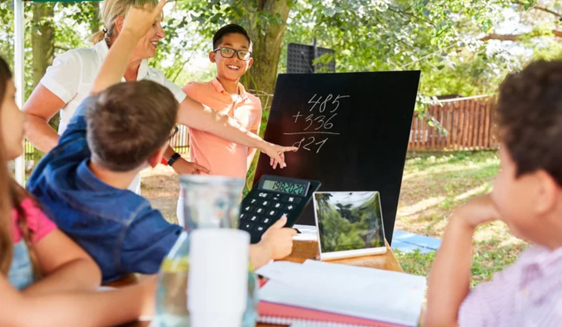 Children collaborating at a table, with a blackboard displaying notes or drawings behind them.