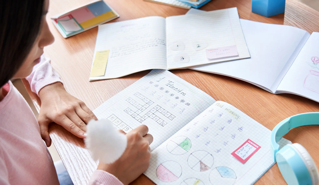 A girl sits at a table, writing in a notebook with a pen, focused on her task.