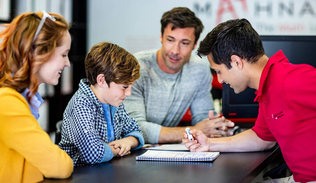 A math tutor engages with a boy and his family at a table, fostering a supportive learning environment.
