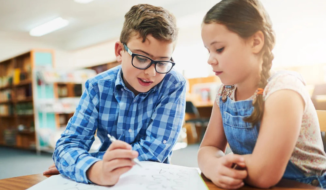 In a library, two children are engaged in writing on paper, surrounded by shelves filled with books.