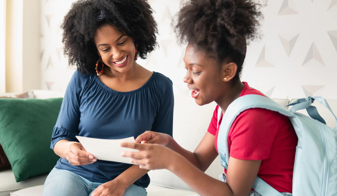 On a couch, a woman and a young girl share a moment, studying a piece of paper with curiosity and engagement.