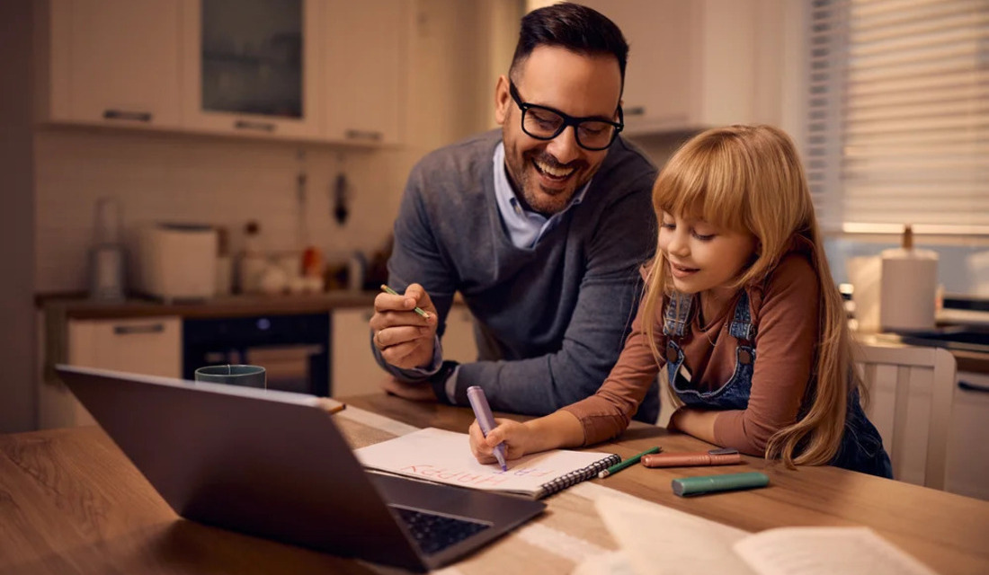 A man assists a young girl with a laptop, both engaged in learning or working in a cozy environment.
