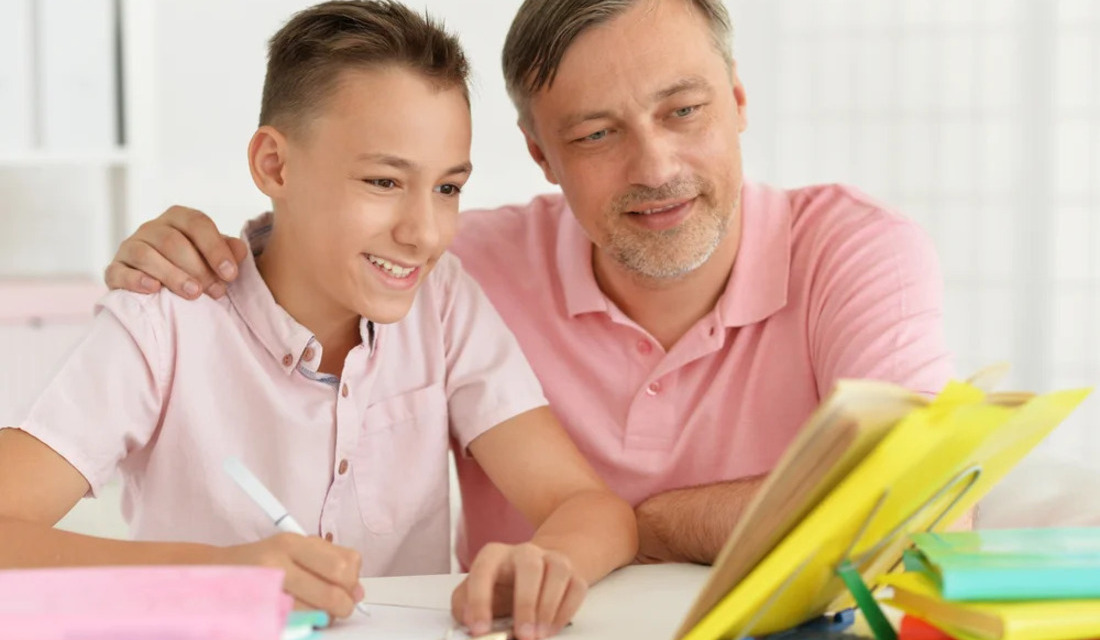 A man and a child share a table, both looking at a book they are reading together.