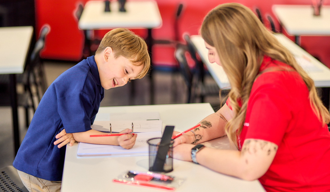 A woman and a child sit at a table, both focused on writing with a pen.