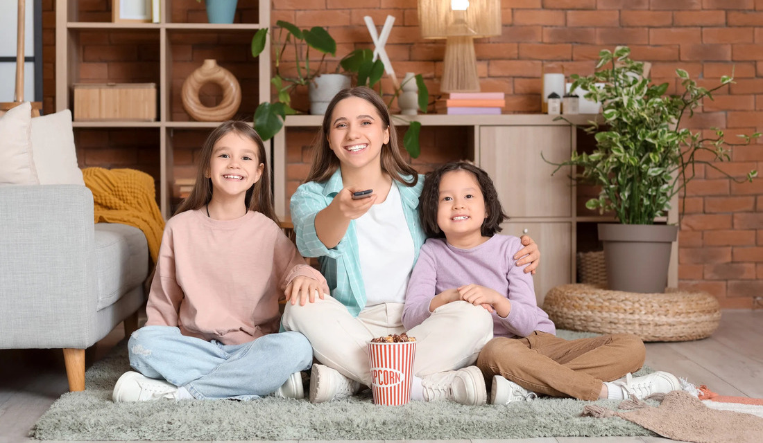 A woman and two children are seated on the floor, enjoying time together against a rustic brick wall backdrop.