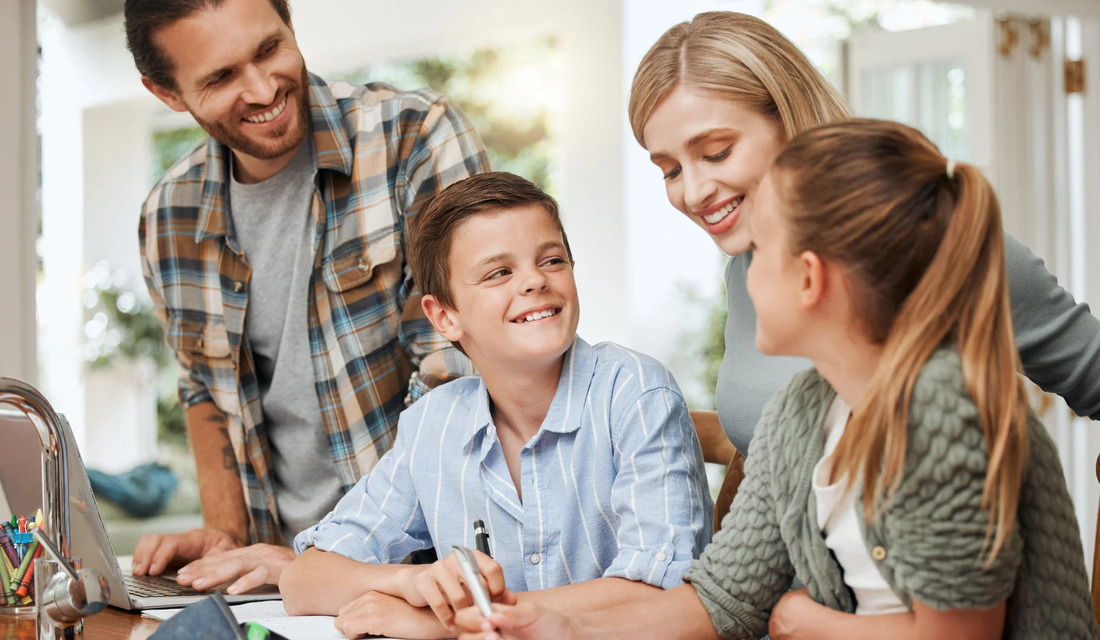 A family of four sits around a table, engaging with a laptop and sharing smiles and conversation.